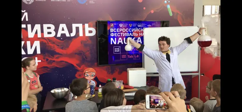 Ведущий в белом халате с поднятыми руками перед детьми на фестивале NAUKA 0+